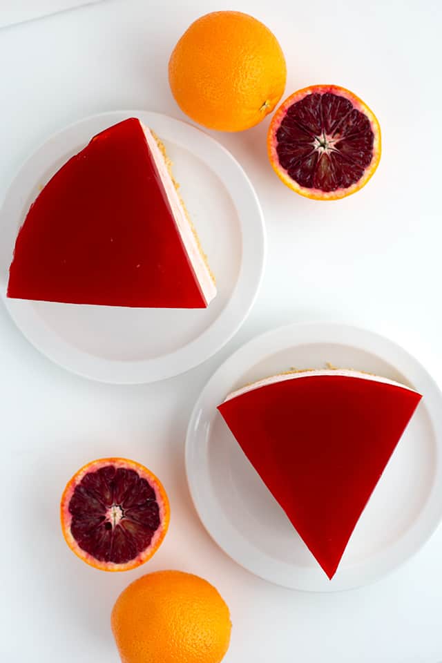 overhead photo of two slices of blood orange cheesescake on white dessert plates with oranges scattered