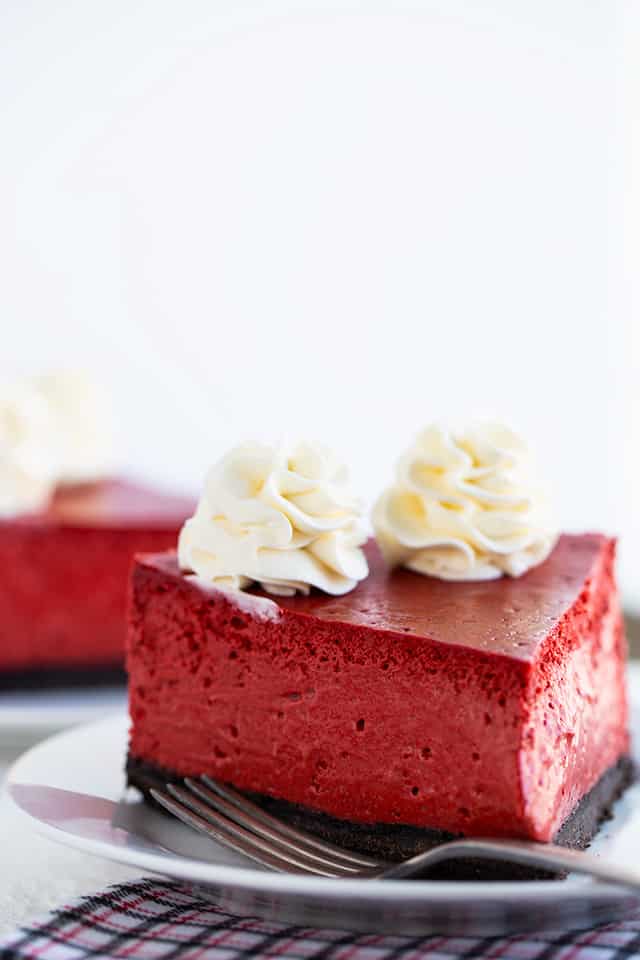 slice of red velvet cheesecake with whipped cream on white dessert plate with fork and a plaid fabric