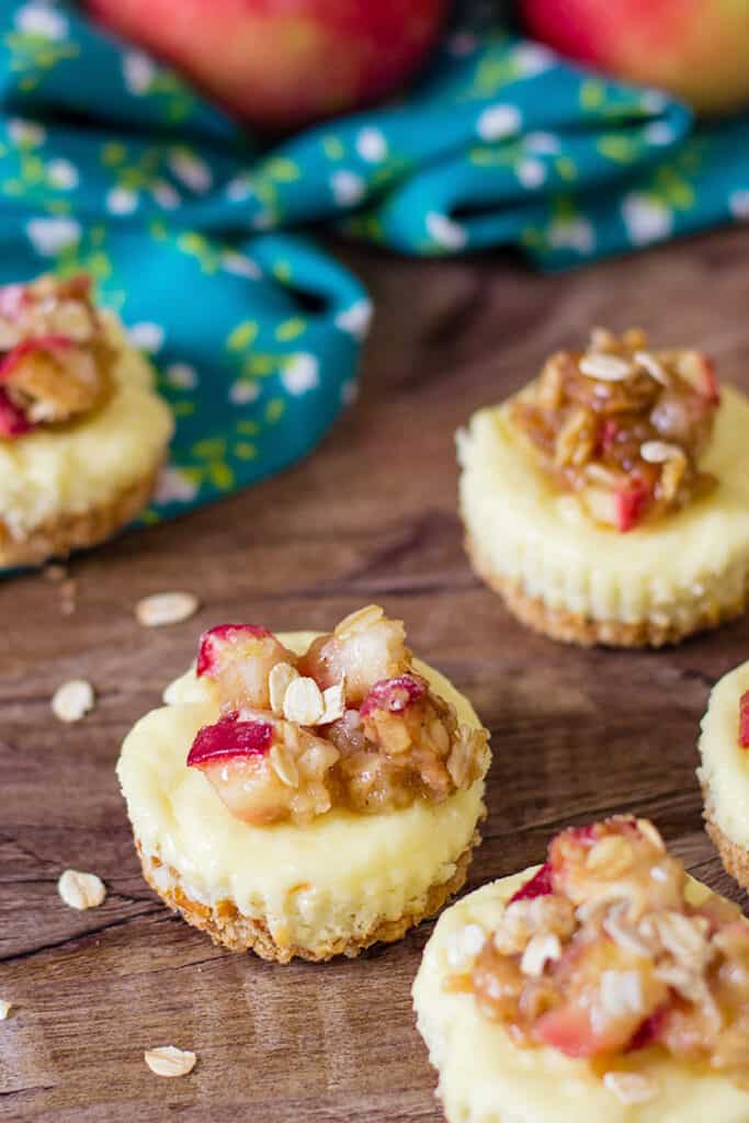 apple crumble cheesecakes on a wood background with oats and other cheesecakes around it and a blue linen behind it