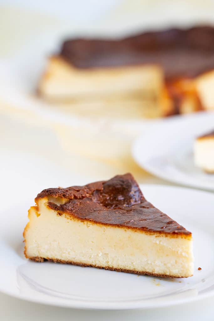 slice of cheesecake on a white plate with the rest of the cheesecake on a dessert platter behind it