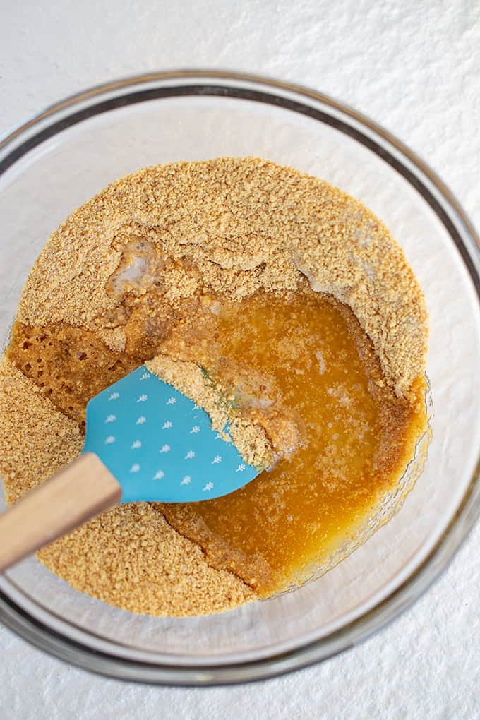 glass bowl with graham cracker crumbs, butter, and a silicone spatula on a white surface